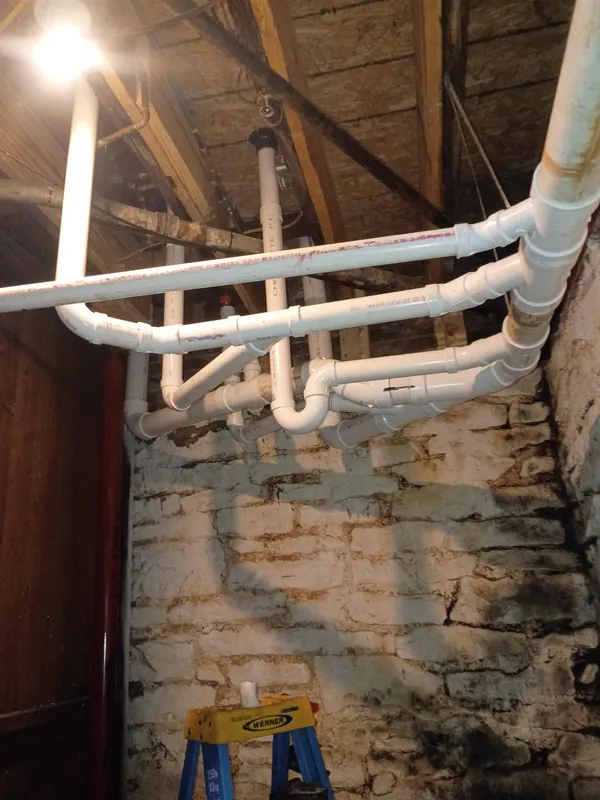 New white PVC drain piping run along the joists and down a stone basement wall