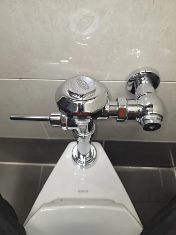 A commercial flush valve above a urinal with a worn handle assembly