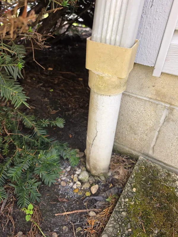 A cracked PVC downspout drain at the base of a house foundation