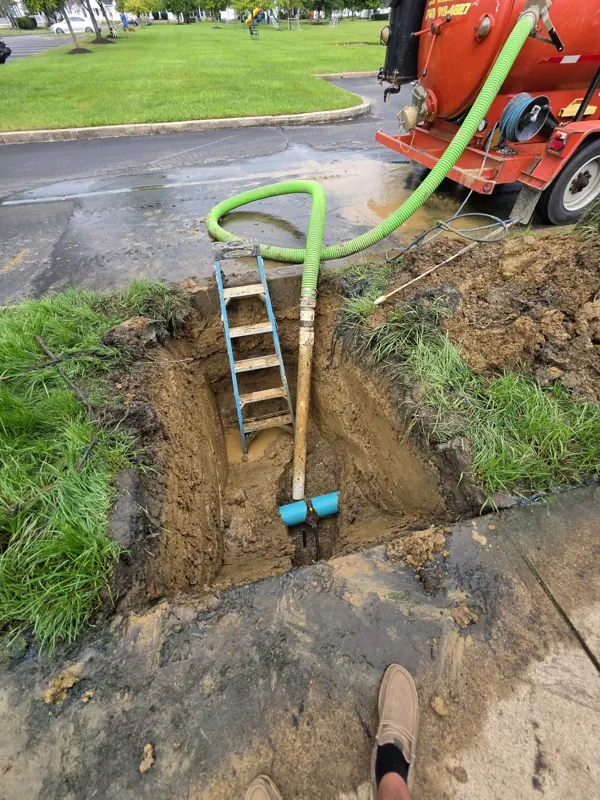 A ladder set into a narrow excavation beside a sidewalk with a suction hose in the hole