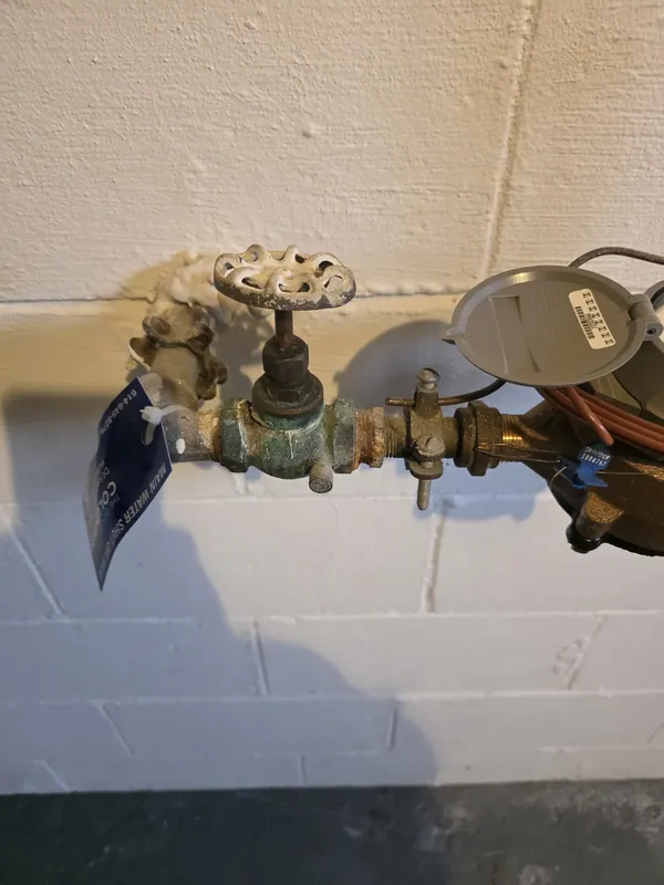 A corroded brass main shut-off valve beside the water meter on a basement block wall